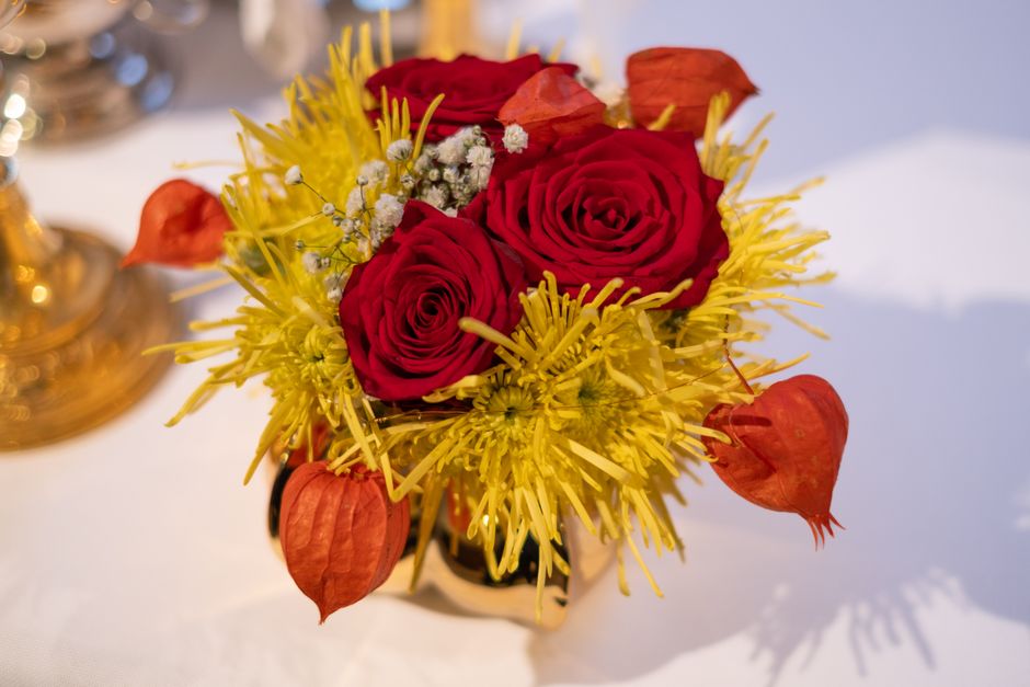 A floral bouquet featuring three large, deep red roses with velvety petals at the center, surrounded by bright yellow spider chrysanthemums with elongated, spiky petals. The arrangement is accented wi