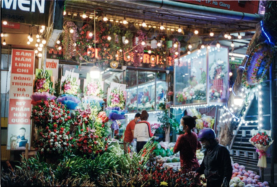 A vibrant flower market scene at night featuring an array of fresh, colorful flowers including pink, red, and white blooms arranged in bouquets and displays. The market stall is decorated with hanging