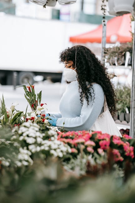A woman with long, curly dark hair wearing a white face mask, light blue sweater, and gloves, is arranging white lilies with red accents at an outdoor flower stall. She stands behind a counter filled 